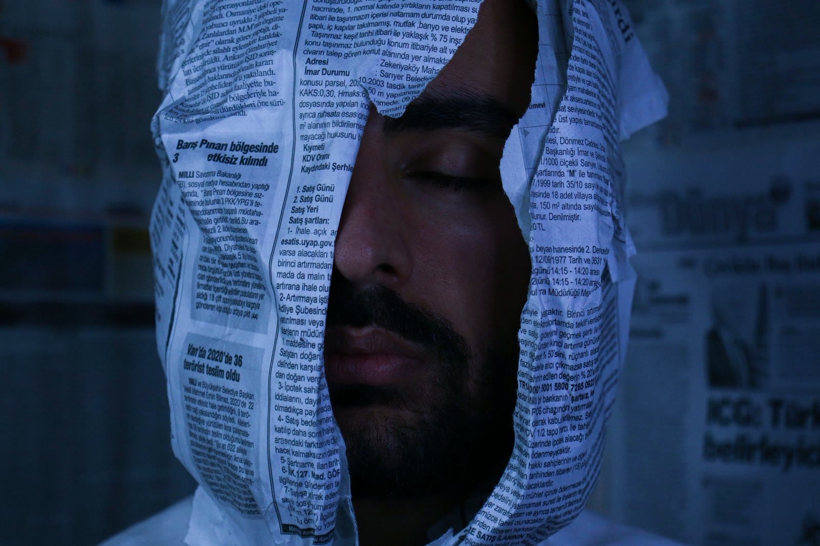 A person with their eyes closed is enveloped in newspaper sheets, creating a surreal or artistic scene. The background also contains newspaper pages, giving a sense of being surrounded by text and pri