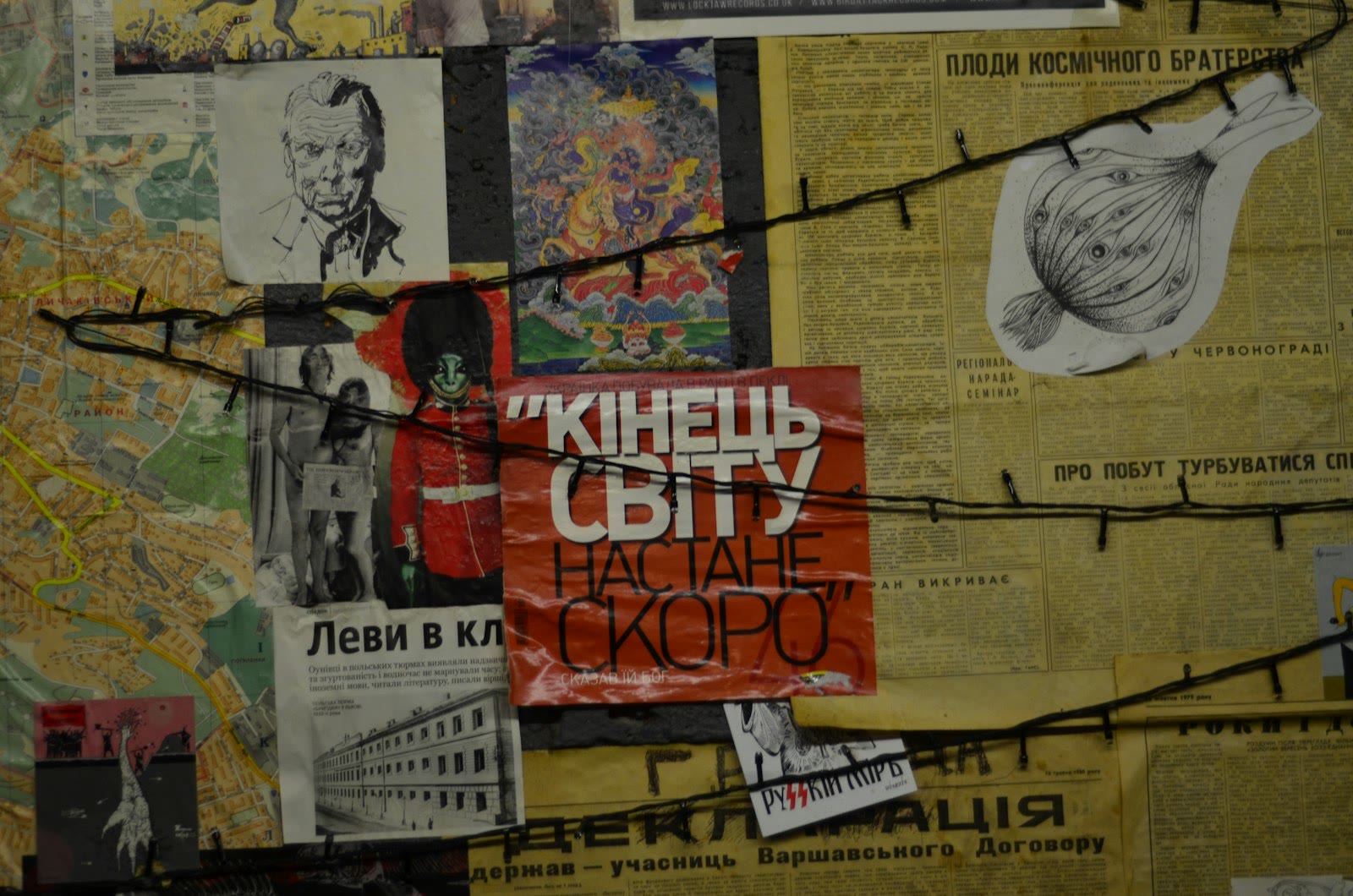 A collage of various newspaper clippings and images, including a large red poster with Cyrillic text, a black and white sketch of a man, and colorful illustrations. Strings run across the collage, add