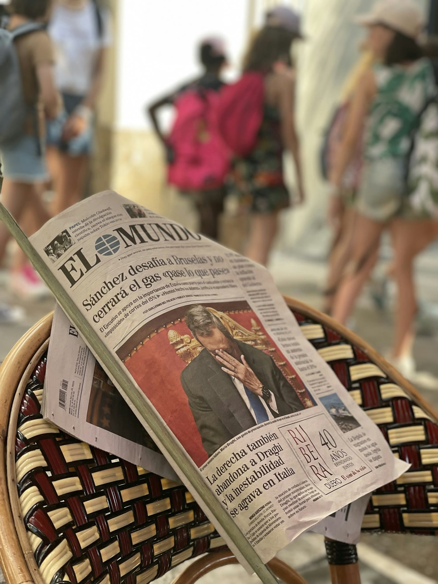 A newspaper is resting on a woven chair, prominently showing a front-page article with a photo of a man in a suit covering his face. The background is blurred, with several people walking, some carryi