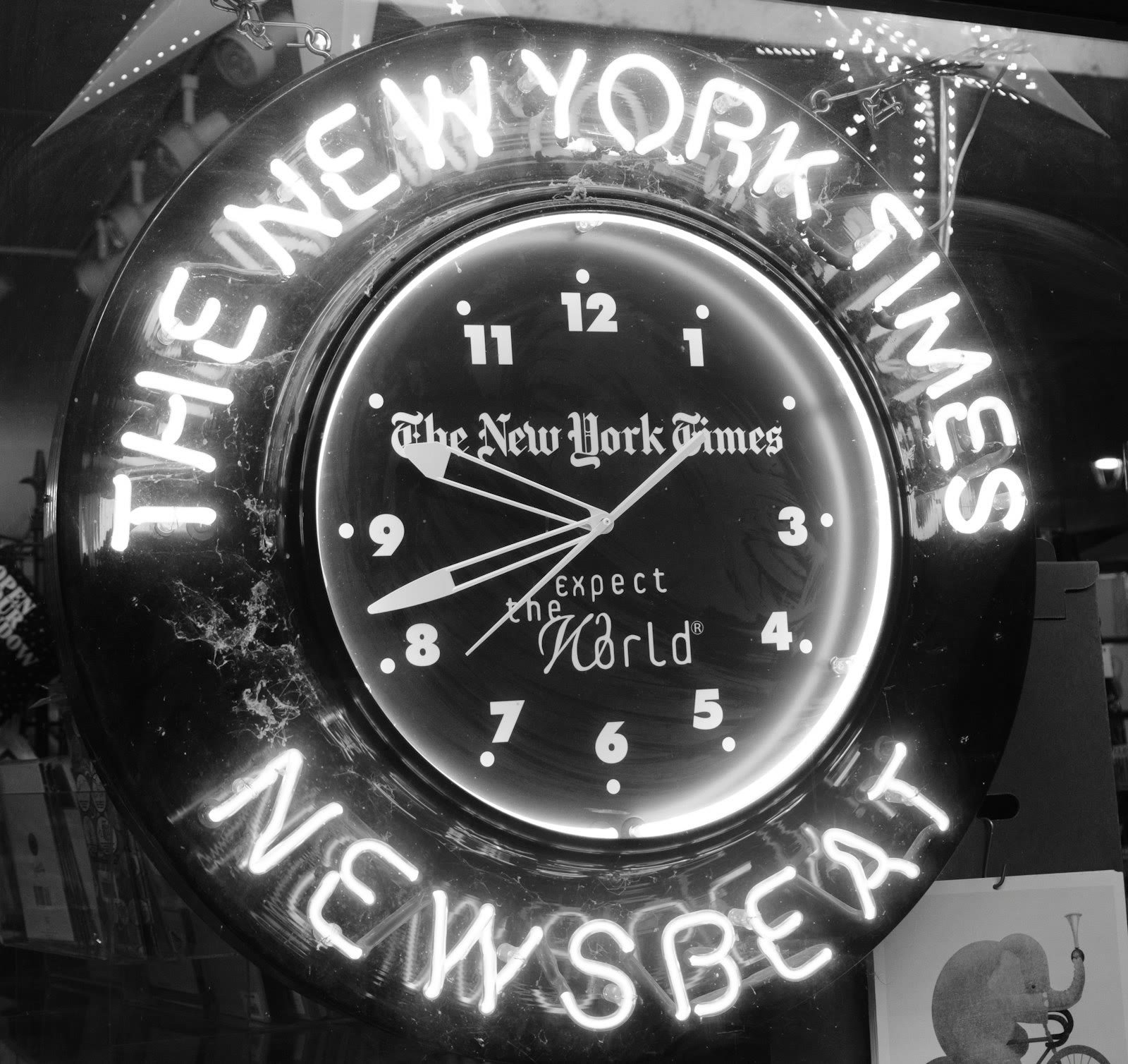 A neon clock with the words 'THE NEW YORK TIMES' and 'NEWSBEAT' illuminated around it. The clock face features the phrase 'expect the World' along with numerals in a classic font. The image is in blac