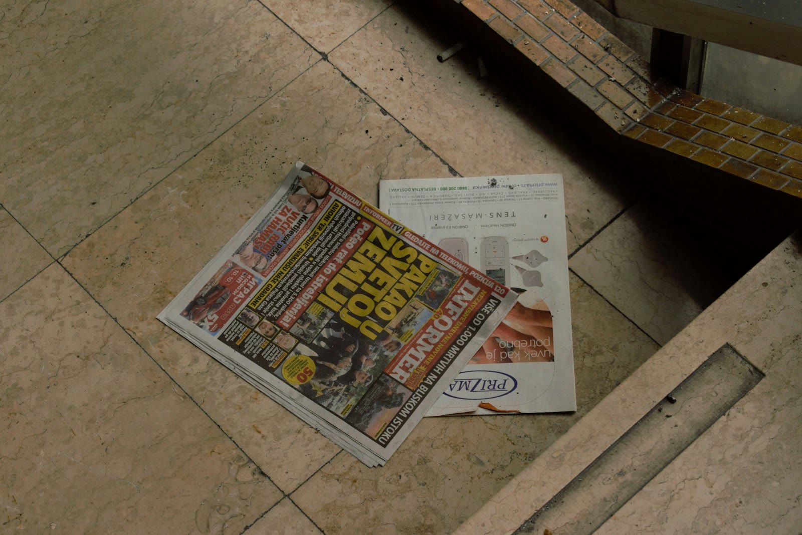 A newspaper with bold headlines and colorful images is placed on a light-colored marble floor. The newspaper features advertisements and various news stories. Adjacent to it is a magazine with an adve