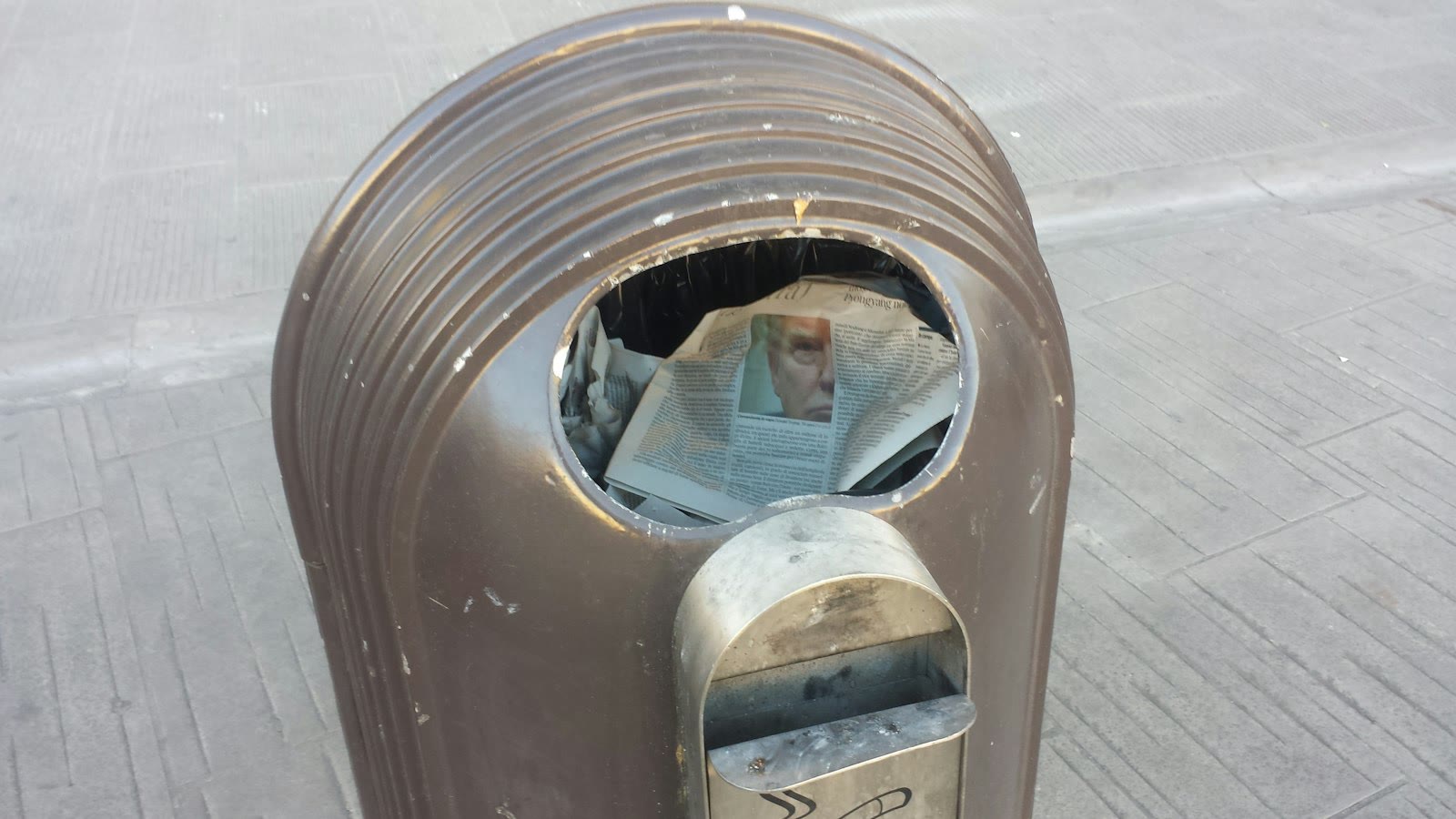 A metallic trash can with a newspaper partially protruding from the opening. The newspaper displays a partially visible image of a face.