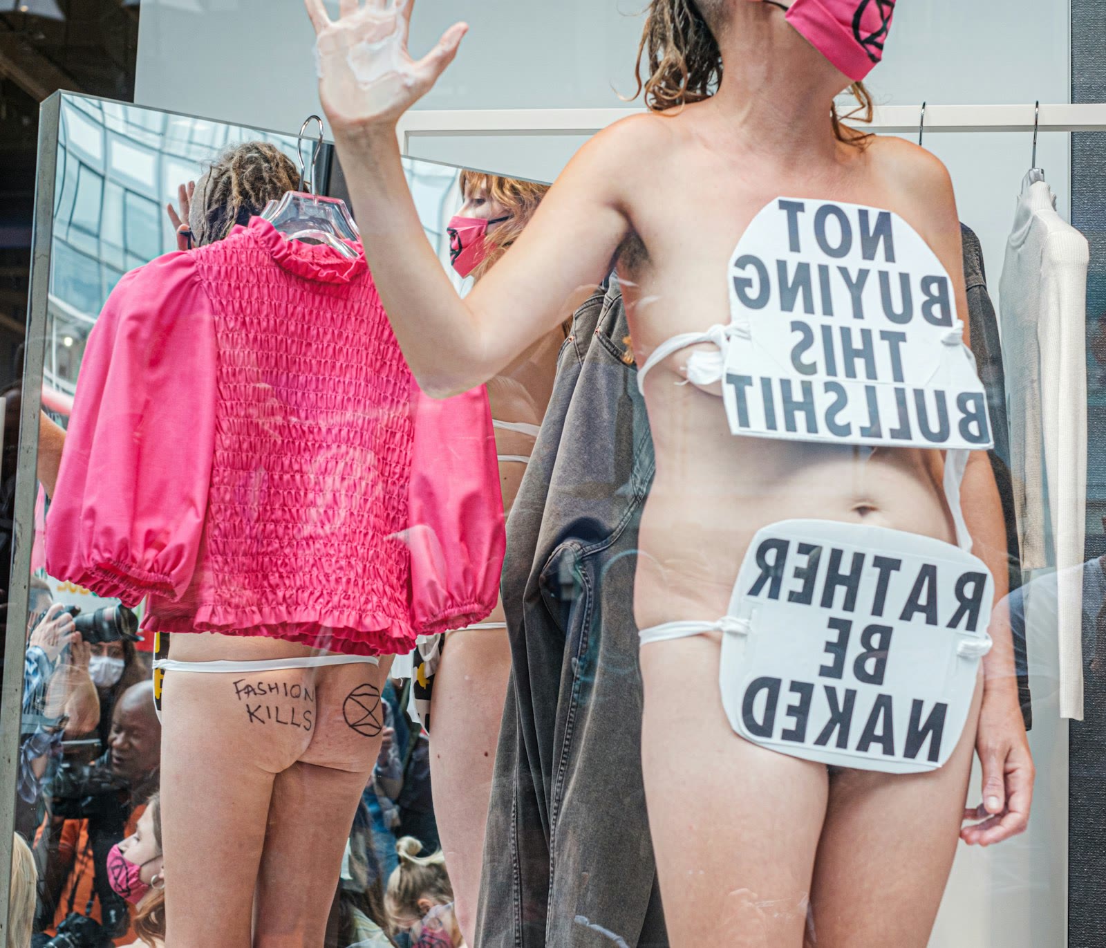 A person stands in a display setting with protest signs that express opposition to consumer culture and fashion. The signs are strategically placed on their body. Reflections in the background show a
