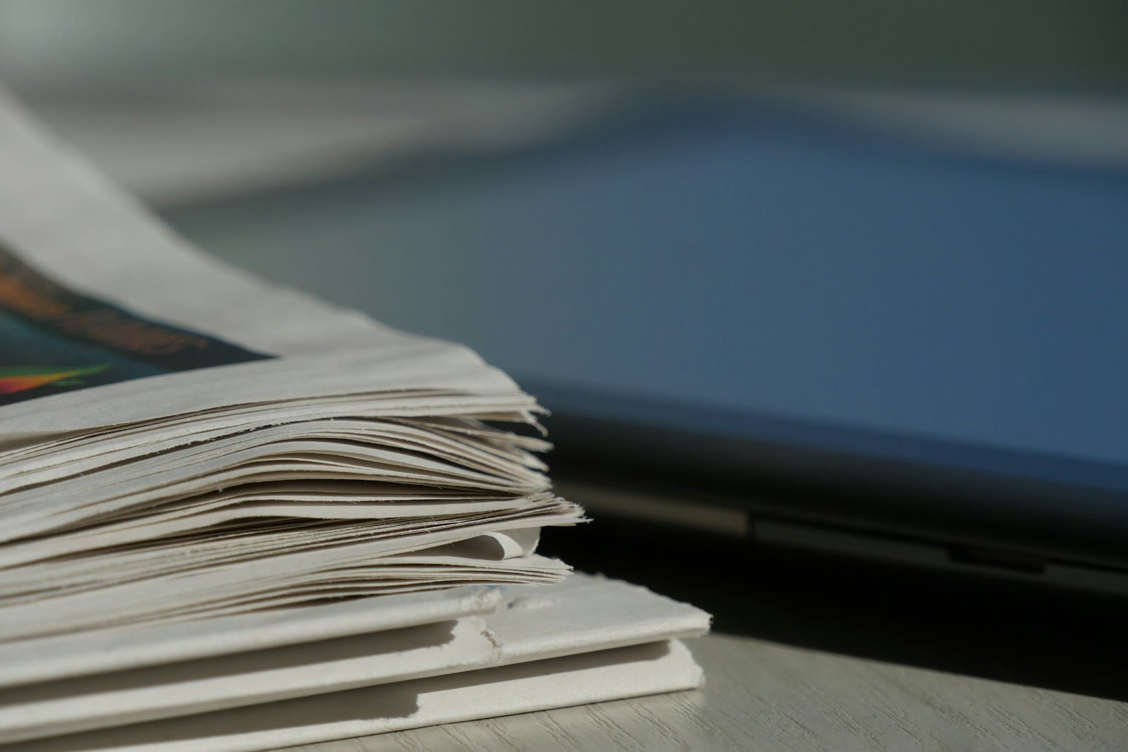 A stack of newspapers lies on a light-colored surface, with some pages slightly curling at the edges. Nearby, part of a laptop or similar electronic device is visible, suggesting a juxtaposition of pr
